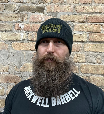 A man wears an black stocking cap with the words "Rockwell Barbell" in gothic letter font on the front of it in army green color. He is also wearing a shirt with the words "Rockwell Barbell" on the front.