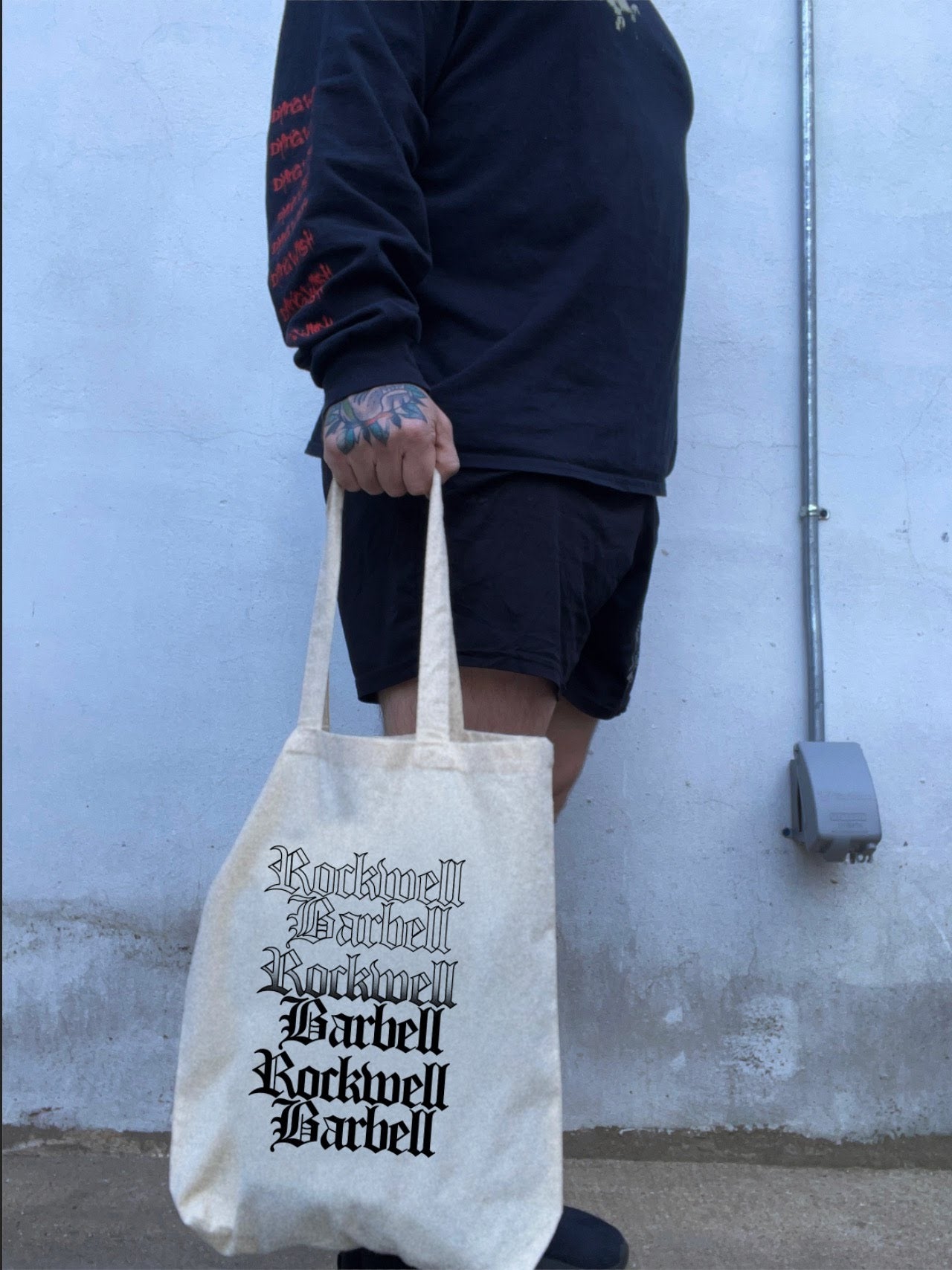 Person holding a tote bag with 'Rockwell Barbell' branding against a plain wall.