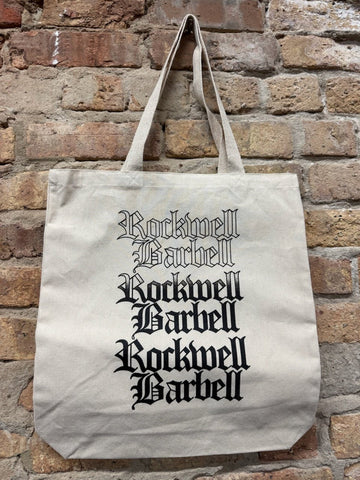 Beige tote bag with 'Rockwell Barbell' text against a brick wall.