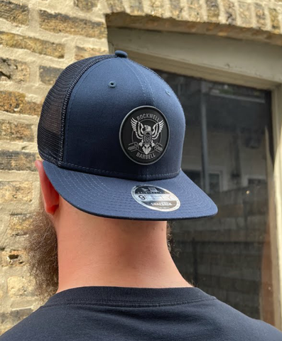 Model qears a navy snapback hat, with the Rockwell Barbell cricle logo in grey and white.