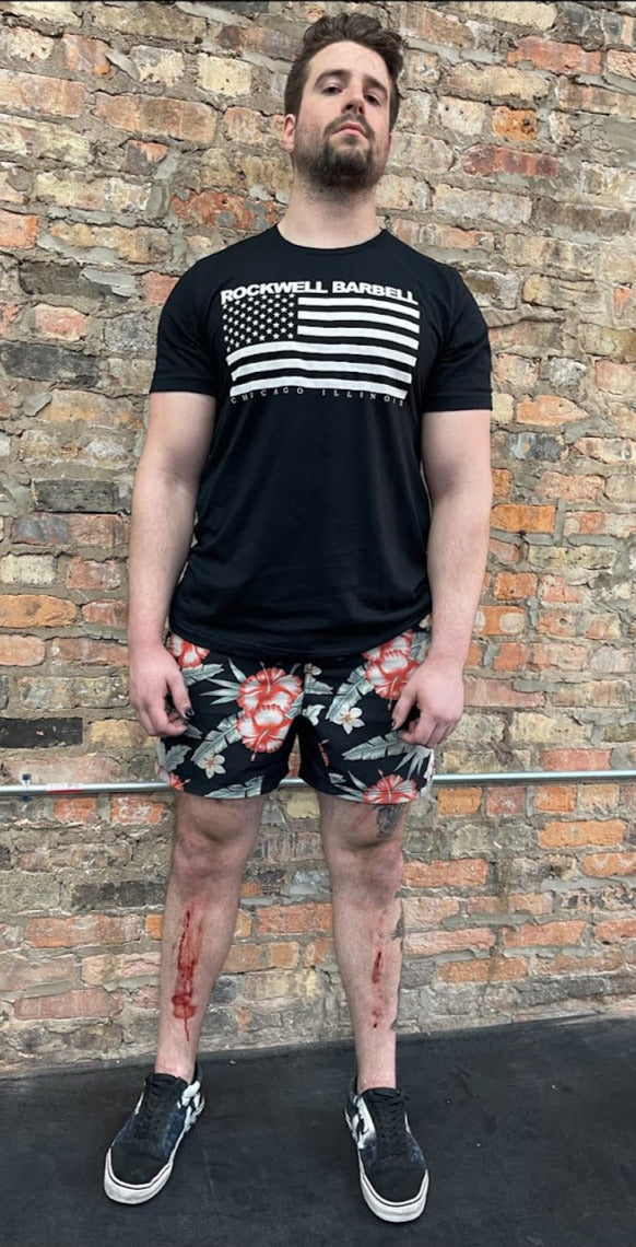 A person wearing a black t-shirt with the words "Rockwell Barbell Chicago, IL" and the USA flag. The design and text is in white, and the t-shirt is black. The person's knees are bloodied slightly..